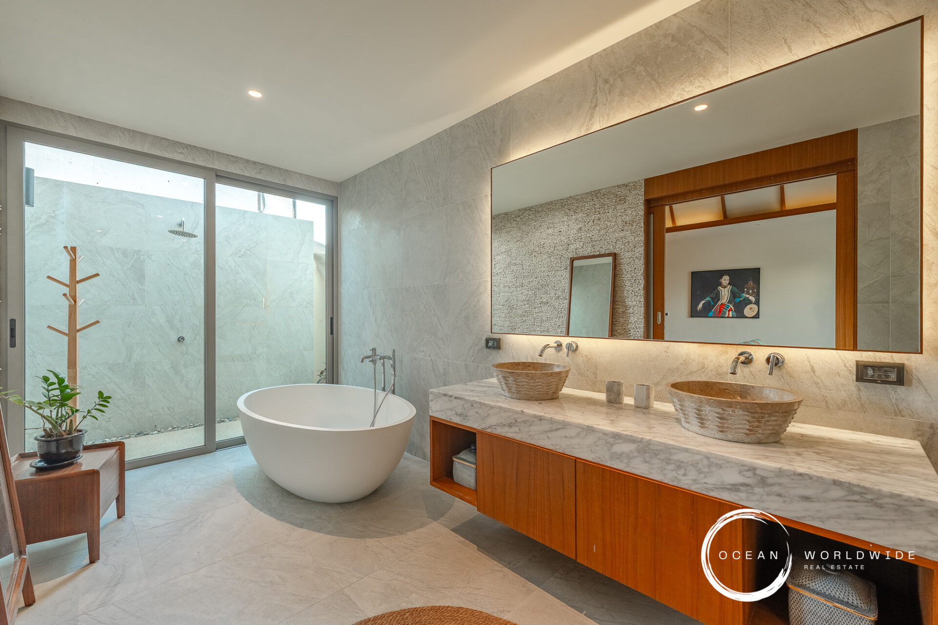 Modern marble bathroom with a freestanding oval bathtub, double vessel sinks, and a large backlit mirror above a wooden vanity.