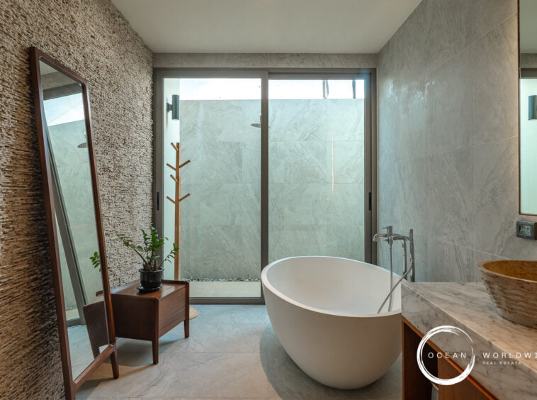 Modern bathroom with a freestanding white tub, marble counters, and a textured stone wall adjacent to a glass door leading to an outdoor shower area.