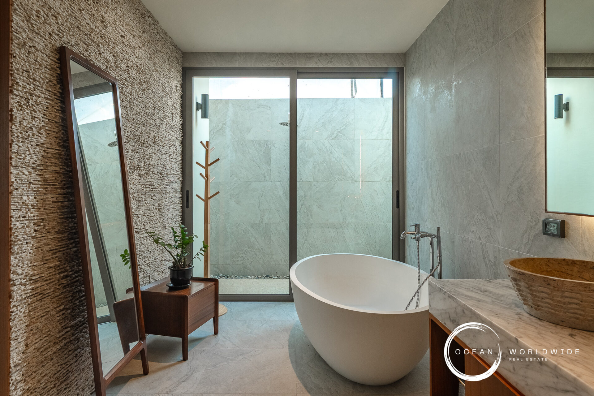 Modern bathroom with a freestanding white tub, marble counters, and a textured stone wall adjacent to a glass door leading to an outdoor shower area.