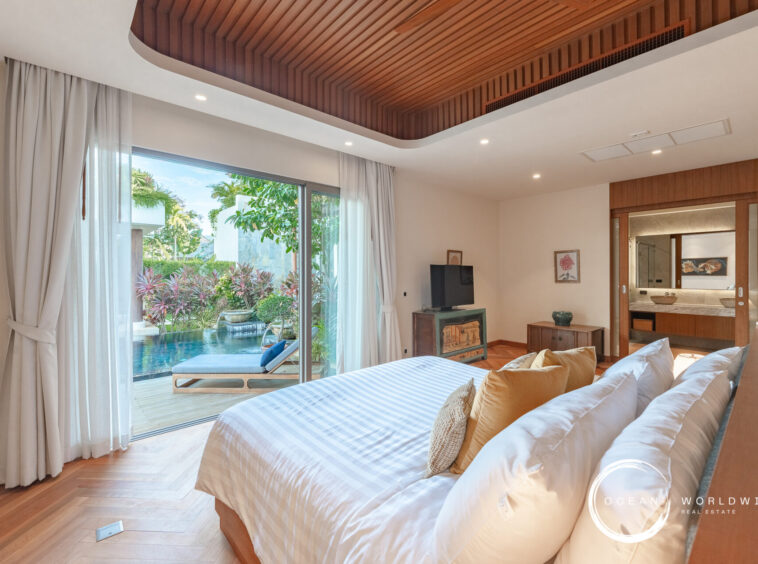 Bright bedroom with a large bed, white linens, and gold pillows, opening to a poolside garden through sliding glass doors with sheer curtains.