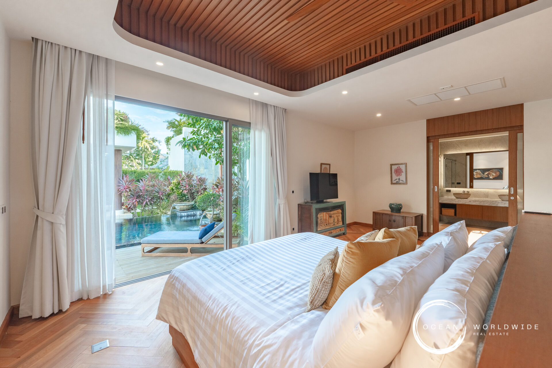 Bright bedroom with a large bed, white linens, and gold pillows, opening to a poolside garden through sliding glass doors with sheer curtains.