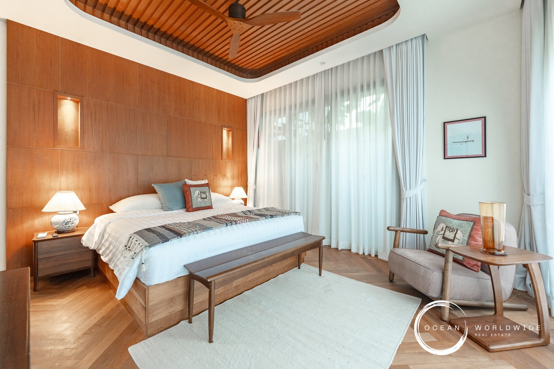 Hotel bedroom with a king bed, wood-paneled headboard, and two bedside lamps. A bench sits at the foot of the bed.