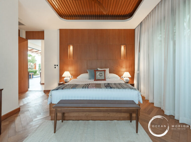 Hotel bedroom with a king bed, tall wooden headboard wall, and two nightstands with lamps on either side. A wooden bench sits at the foot of the bed, and sheer white-curtained windows run along the right side.