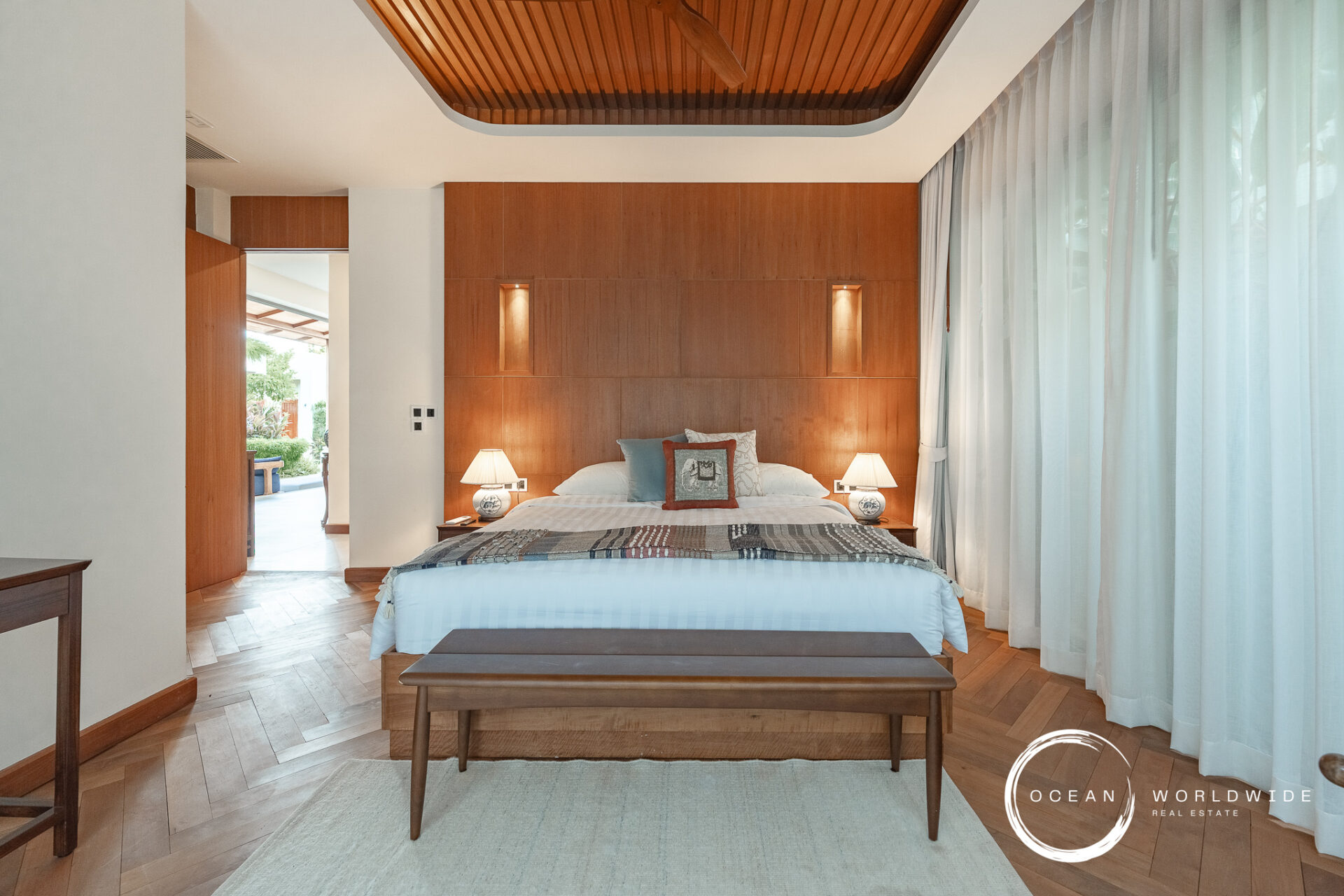 Hotel bedroom with a king bed, tall wooden headboard wall, and two nightstands with lamps on either side. A wooden bench sits at the foot of the bed, and sheer white-curtained windows run along the right side.