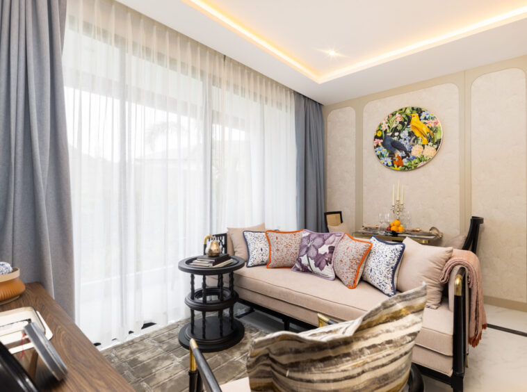 Bright living room with sheer white curtains, a beige sofa with colorful patterned cushions, and a round nature-themed wall art piece in the corner.