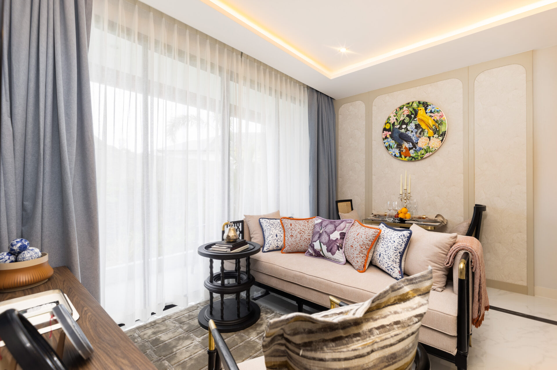 Bright living room with sheer white curtains, a beige sofa with colorful patterned cushions, and a round nature-themed wall art piece in the corner.