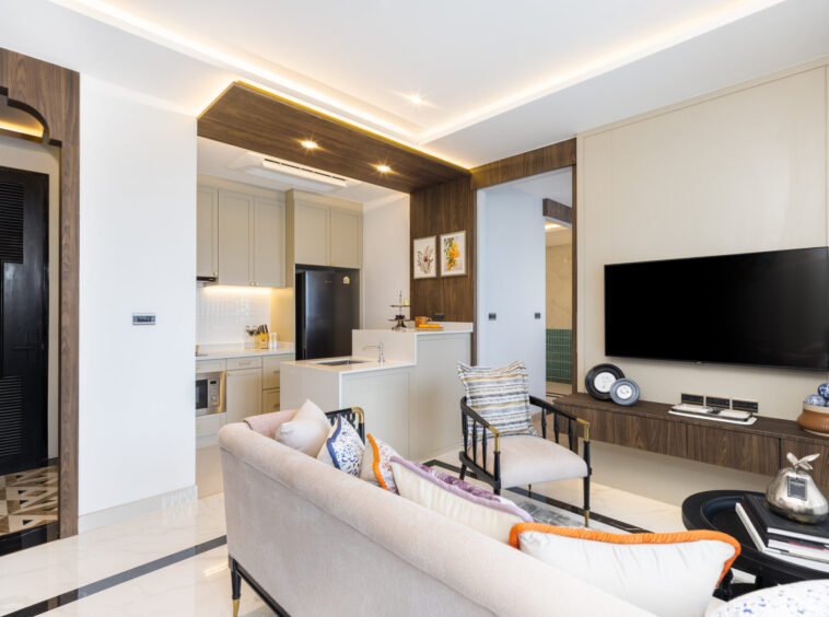 Open-plan living room with beige sofa facing a wall-mounted TV and a wooden console, modern kitchen to the left, and decorative cushions.