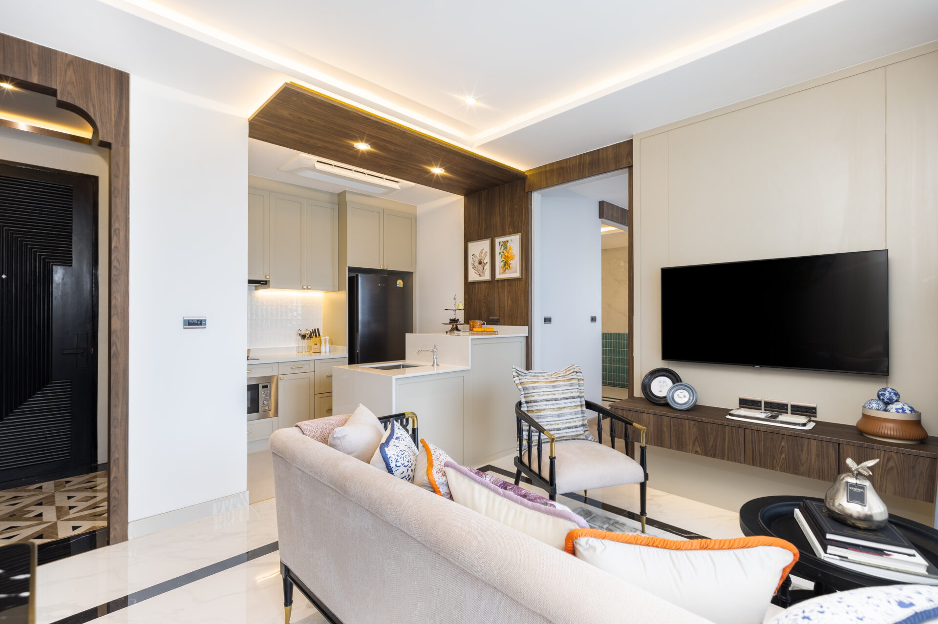 Open-plan living room with beige sofa facing a wall-mounted TV and a wooden console, modern kitchen to the left, and decorative cushions.