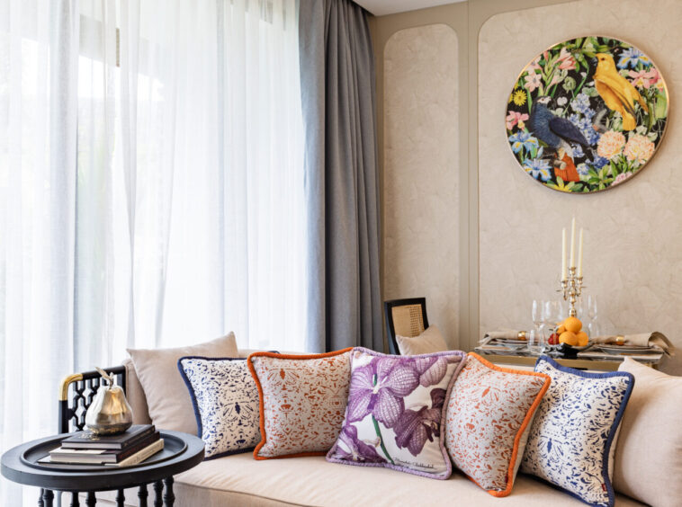 Cozy living room corner with a beige sofa, colorful patterned cushions, sheer white curtains, and a round floral bird painting.