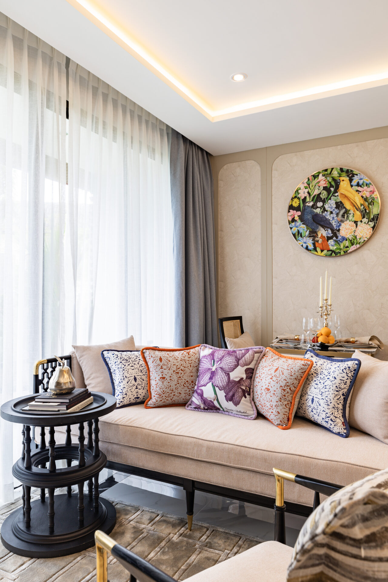 Cozy living room corner with a beige sofa, colorful patterned cushions, sheer white curtains, and a round floral bird painting.