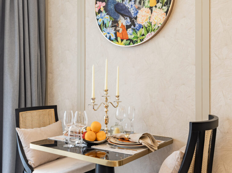 Elegant dining nook with a colorful round bird painting on a beige patterned wall above a table set with candles and fruit centerpiece.