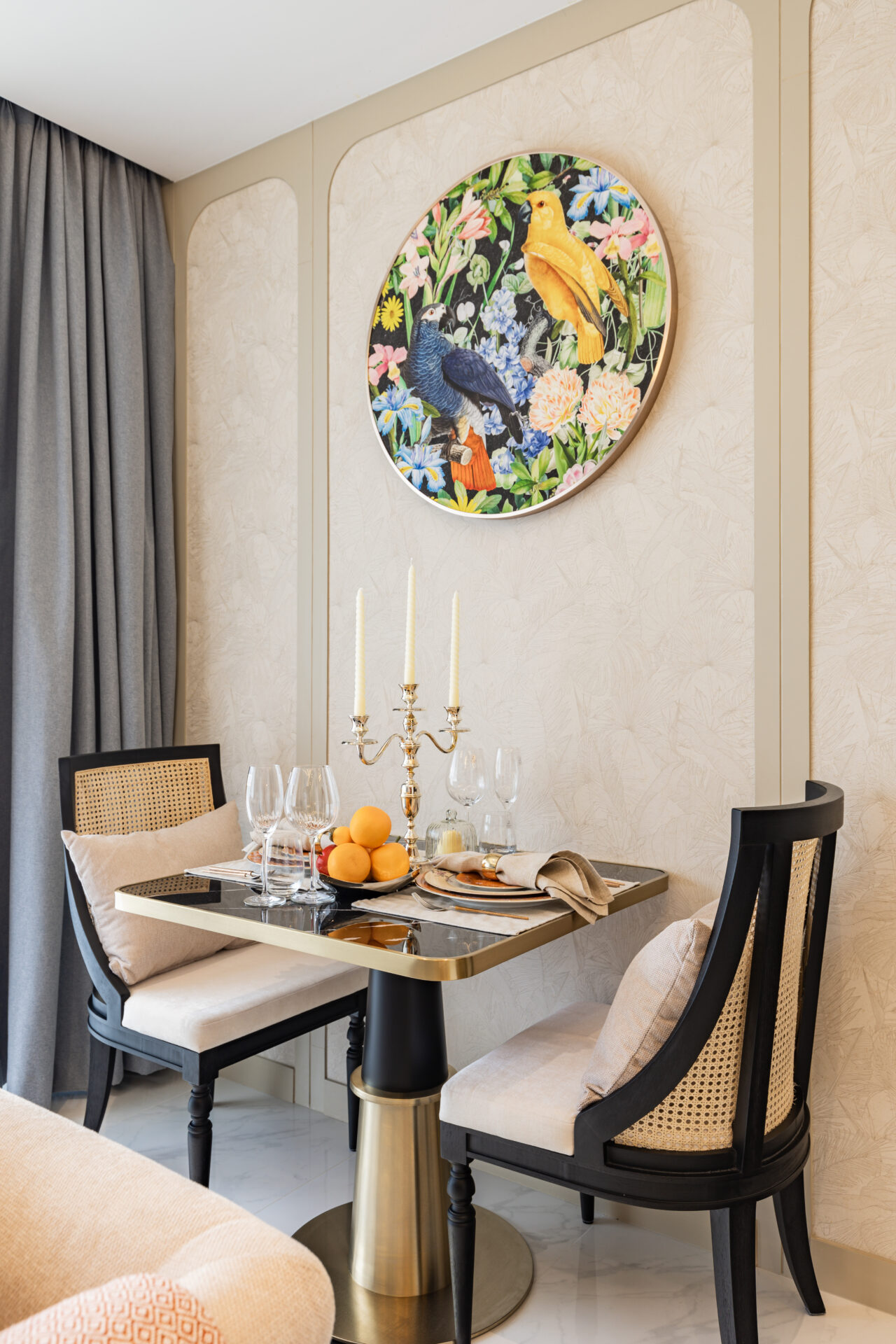Elegant dining nook with a colorful round bird painting on a beige patterned wall above a table set with candles and fruit centerpiece.