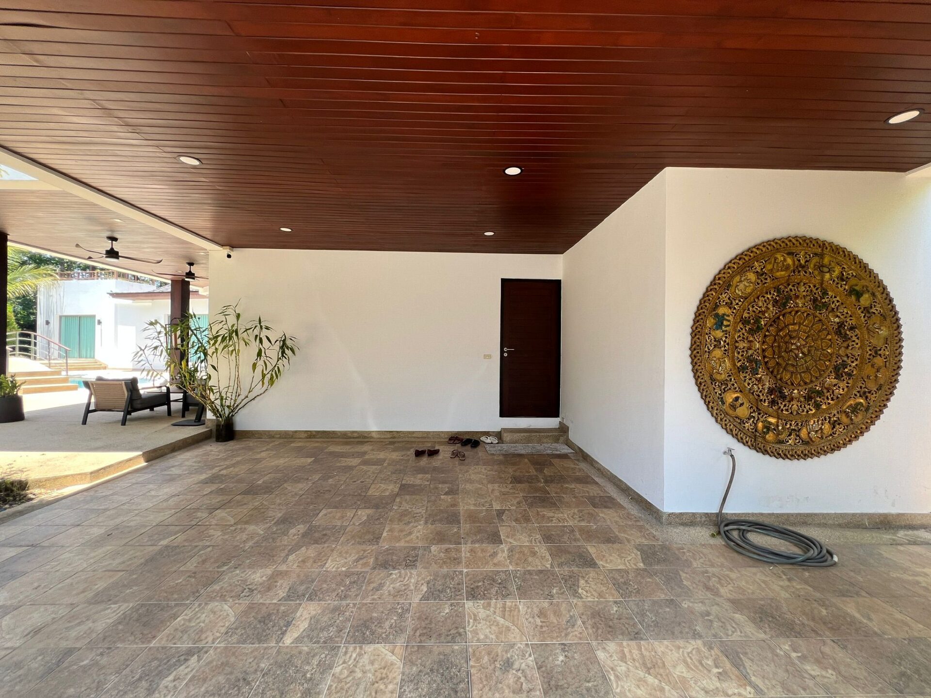 Covered outdoor patio with a wood-paneled ceiling, white walls, and a large ornate gold circular wall art (mandala).