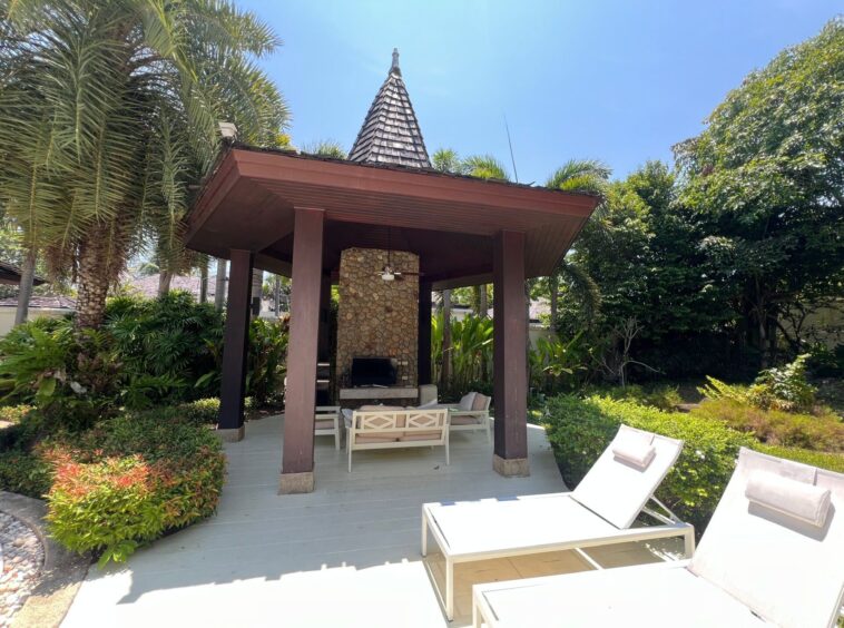 Outdoor pavilion with a stone fireplace and beige seating, surrounded by palm trees and tropical plants in a sunny backyard.