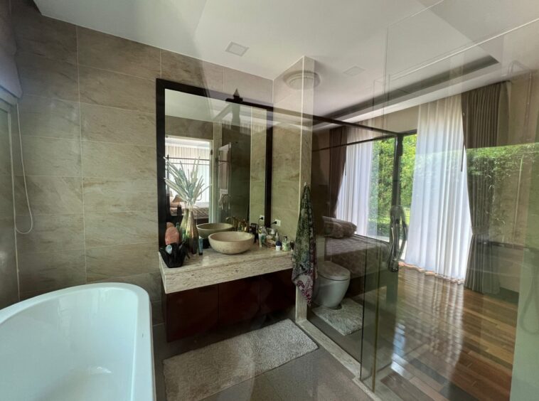 Modern bathroom with glass shower enclosure, double sink vanity, and freestanding tub; large mirror reflects greenery outside.