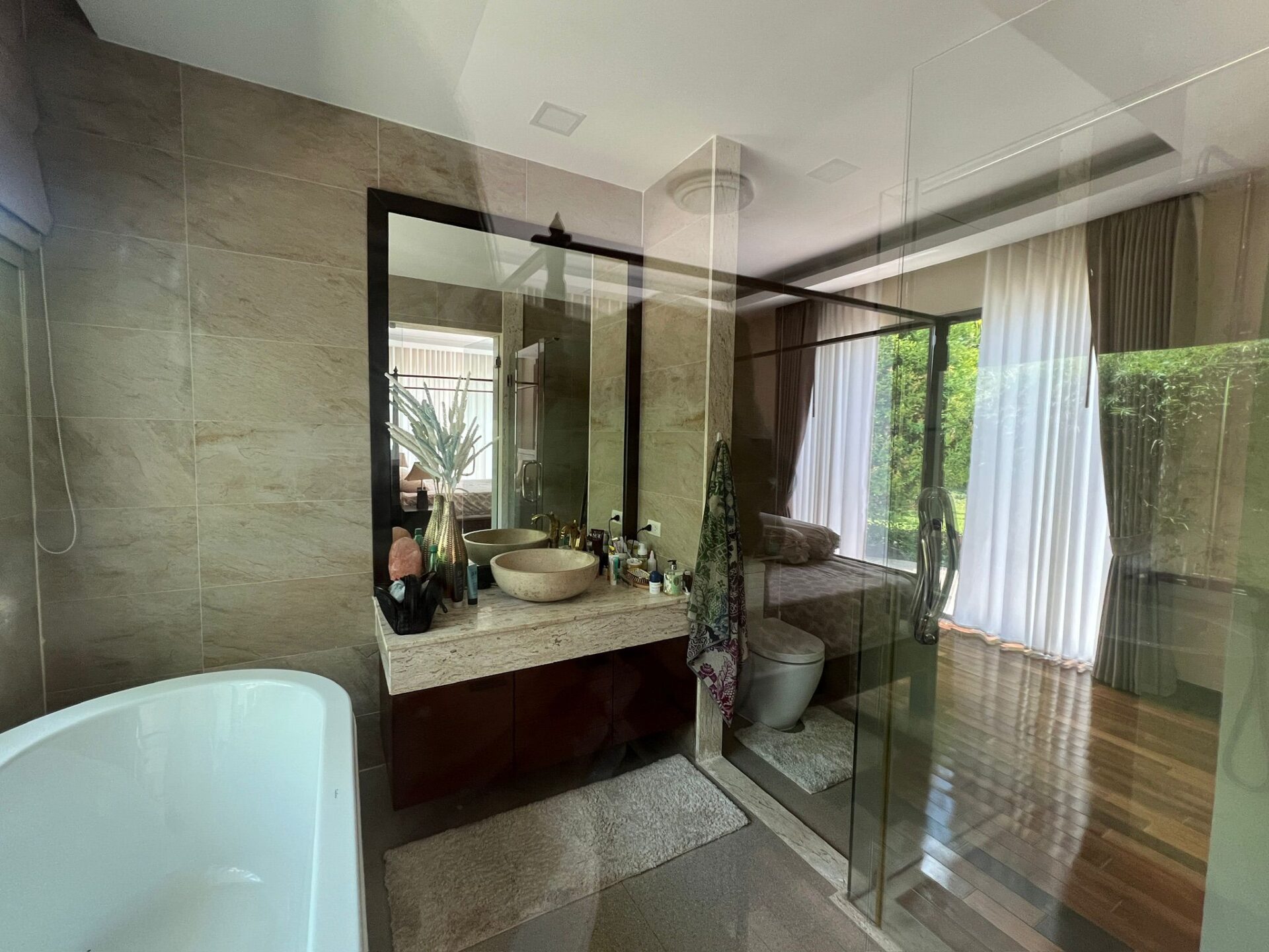 Modern bathroom with glass shower enclosure, double sink vanity, and freestanding tub; large mirror reflects greenery outside.