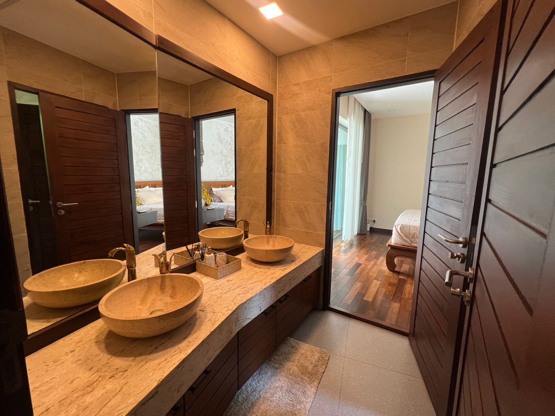 Double-sink bathroom vanity with beige stone bowls and gold fixtures on a marble countertop, mirrored wall, and dark wood cabinets.