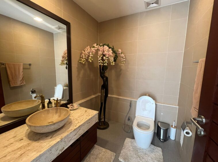 Modern bathroom with a double marble sink vanity, large mirror, and a toilet, accented by a tall floral arrangement.
