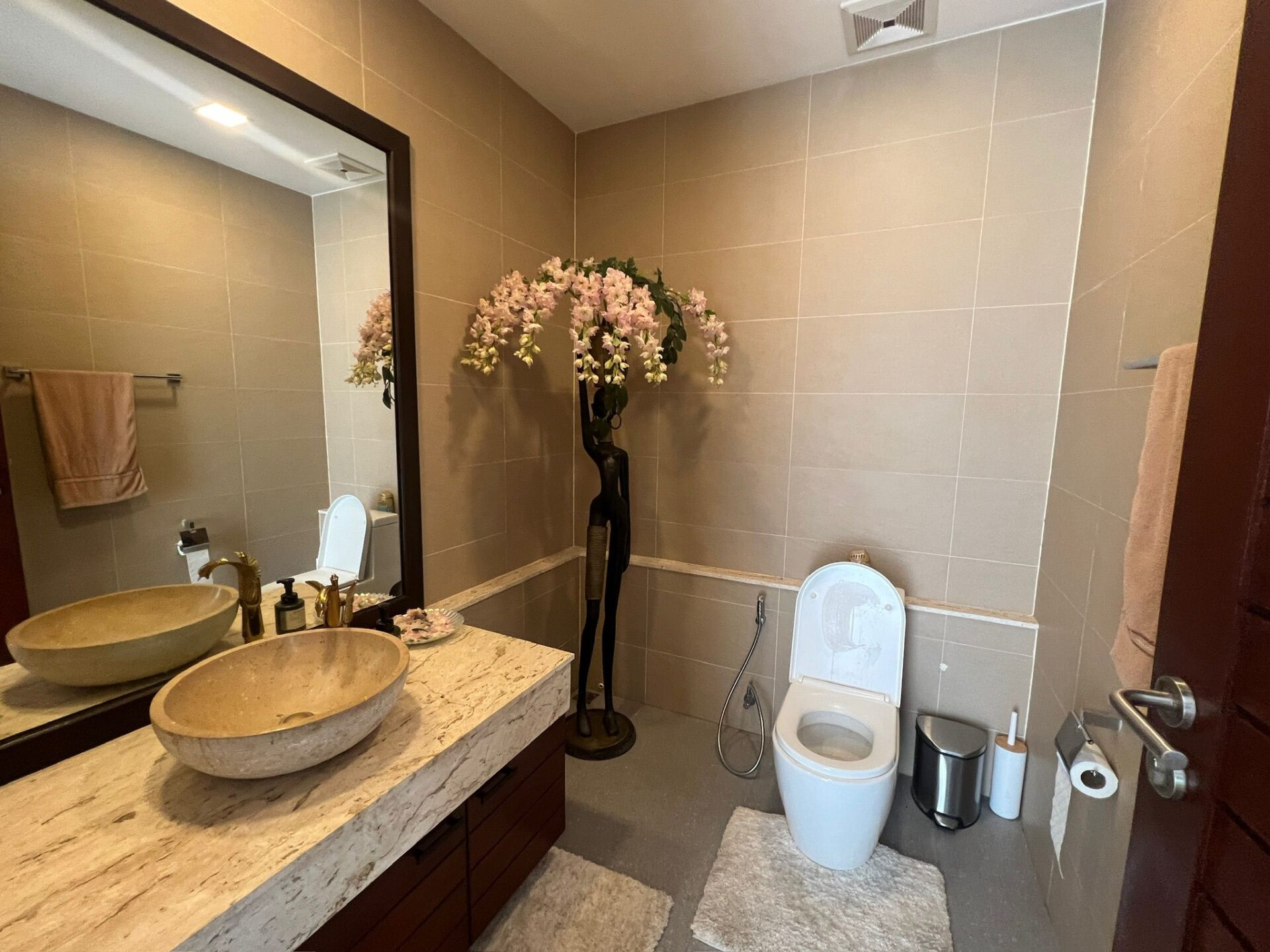 Modern bathroom with a double marble sink vanity, large mirror, and a toilet, accented by a tall floral arrangement.