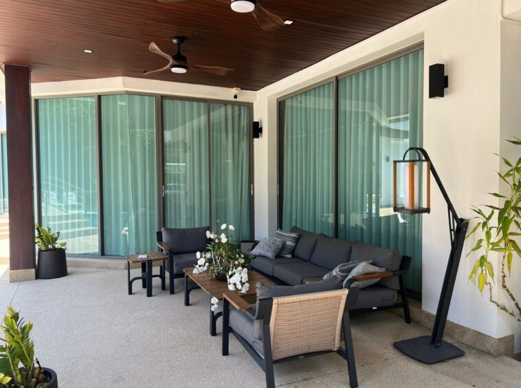 Outdoor covered patio with a dark wicker sofa set, glass sliding doors, and potted plants; cream curtains inside, wooden ceiling, and a hanging lantern.