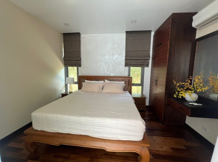 Bedroom with a white bed, wooden headboard, and matching nightstands; brown Roman shades over two windows behind the bed.