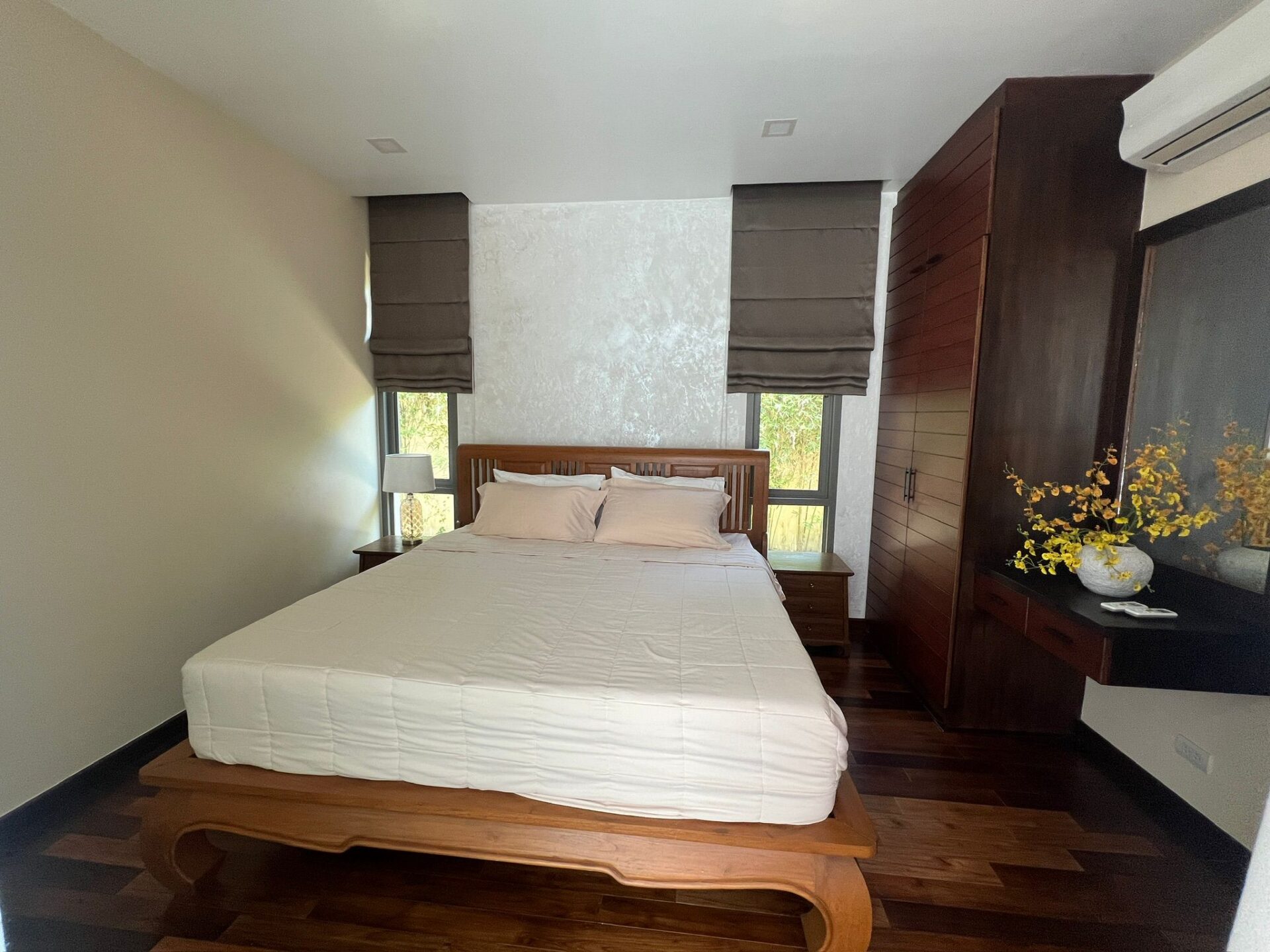 Bedroom with a white bed, wooden headboard, and matching nightstands; brown Roman shades over two windows behind the bed.
