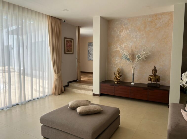 Living room interior with sheer white curtains, beige drapes, and a dark wood console displaying Buddha statues and a tall vase of dried grasses on a textured wall.