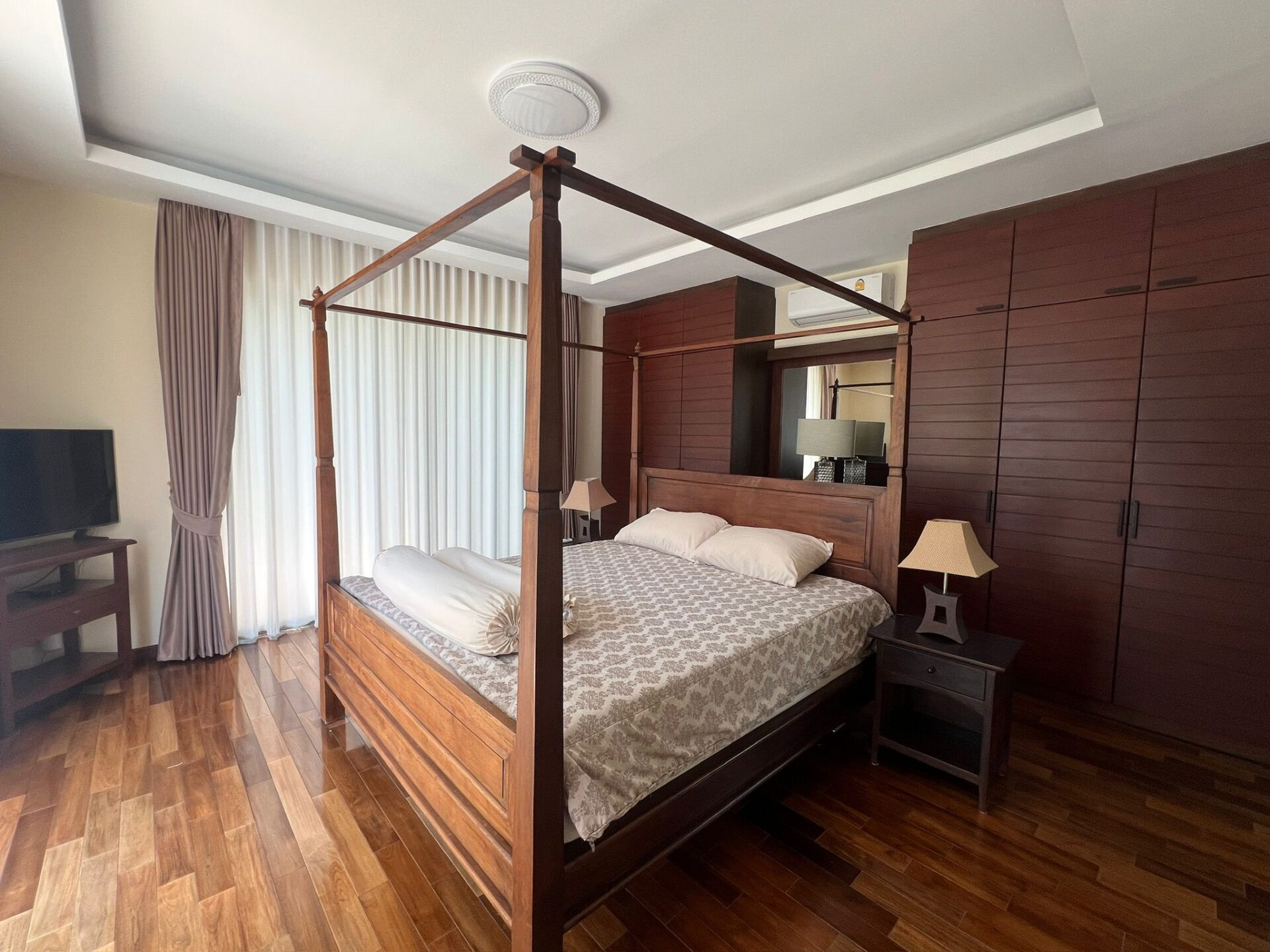 Wood four-poster bed in a brown-toned bedroom with matching nightstands and built-in wardrobes, plus sheer white curtains.