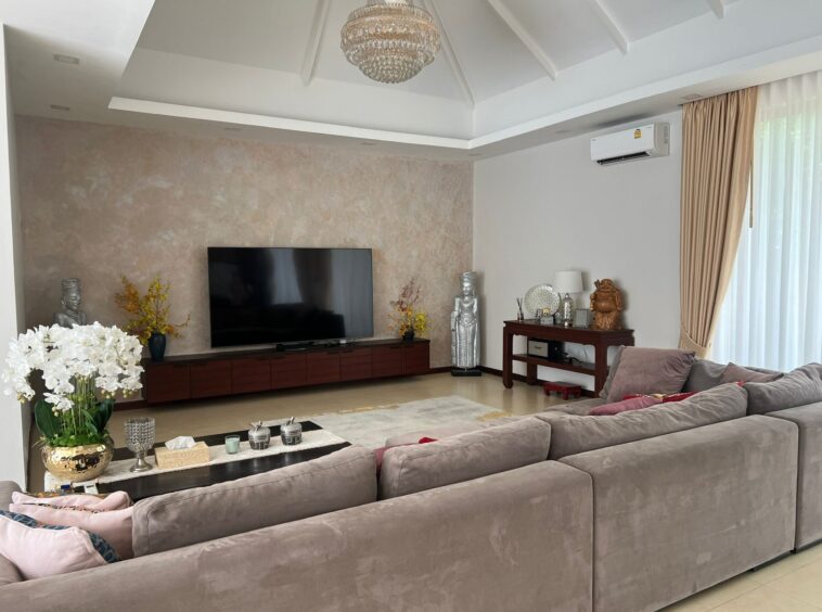 Living room with a large gray sectional sofa facing a wall-mounted flat-screen TV, beige accent wall, and a chandelier overhead.