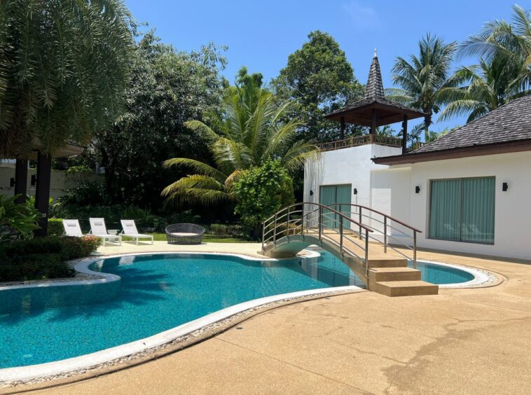 Poolside view of a blue, curved swimming pool with lounge chairs, palm trees, and a wooden gazebo on a raised deck beside a white building.
