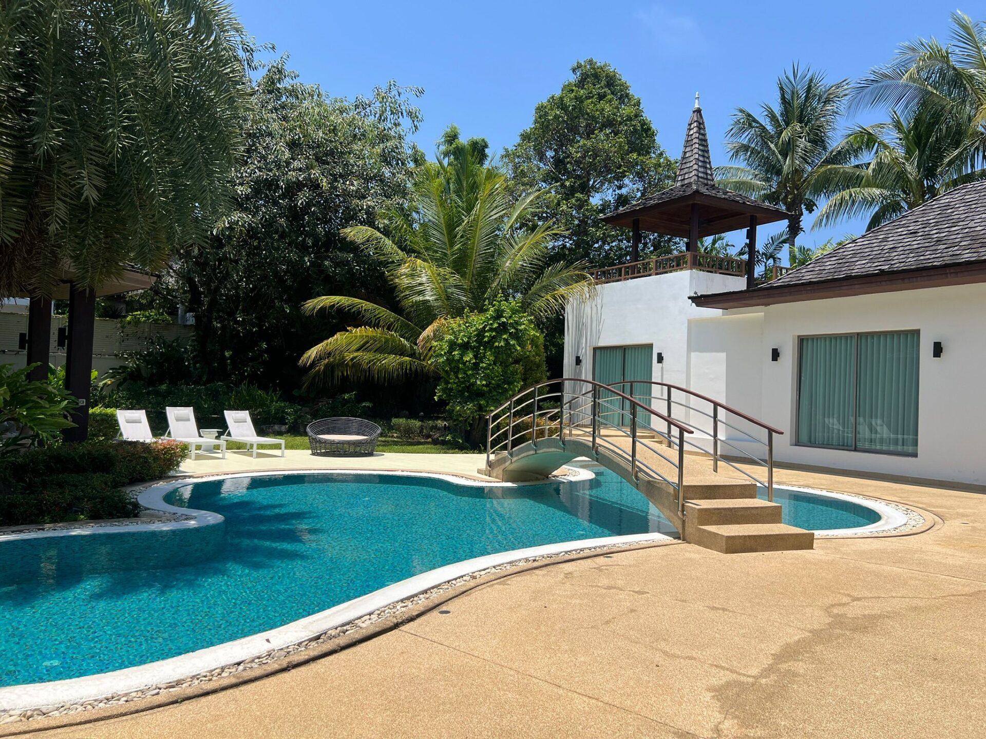 Poolside view of a blue, curved swimming pool with lounge chairs, palm trees, and a wooden gazebo on a raised deck beside a white building.