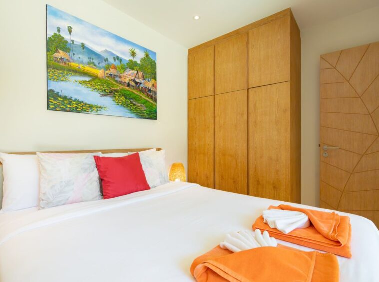 Bright hotel-style bedroom with a large bed, white linens, and orange towels folded at the foot; wooden wardrobes and a geometric-pattern door to the right; a colorful village painting hangs above the bed.