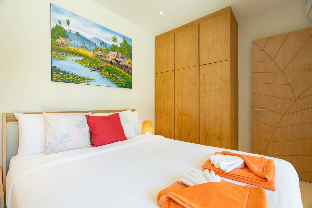 Bright hotel-style bedroom with a large bed, white linens, and orange towels folded at the foot; wooden wardrobes and a geometric-pattern door to the right; a colorful village painting hangs above the bed.