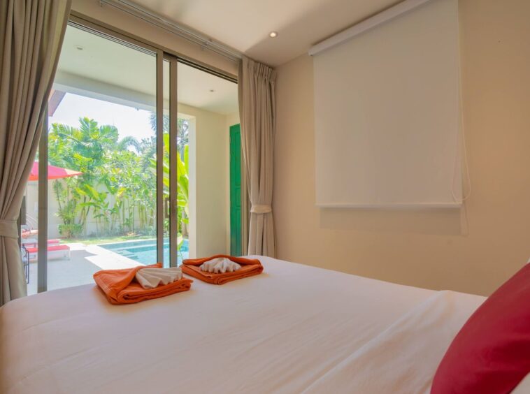 Hotel bedroom with a sliding glass door to a pool deck, white bedding, and orange folded towels on the bed.