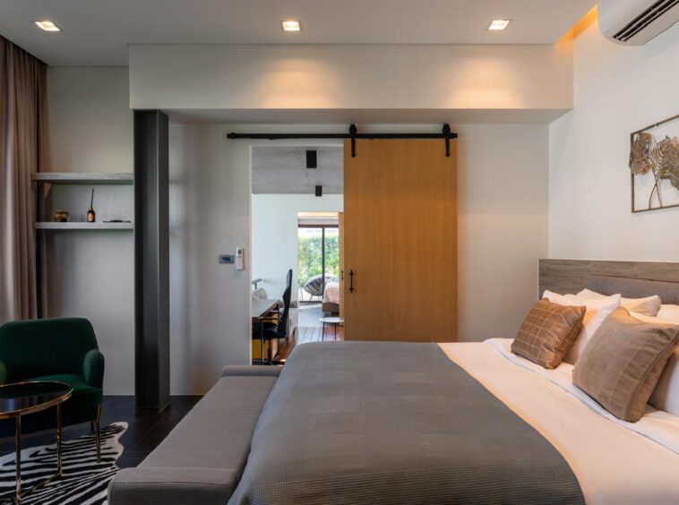 Modern hotel bedroom with a large bed, gray bedding and beige cushions, and a wooden sliding barn door open to a seating area.