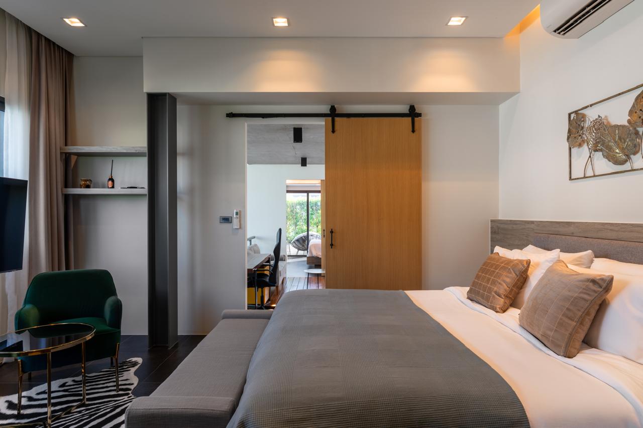 Modern hotel bedroom with a large bed, gray bedding and beige cushions, and a wooden sliding barn door open to a seating area.