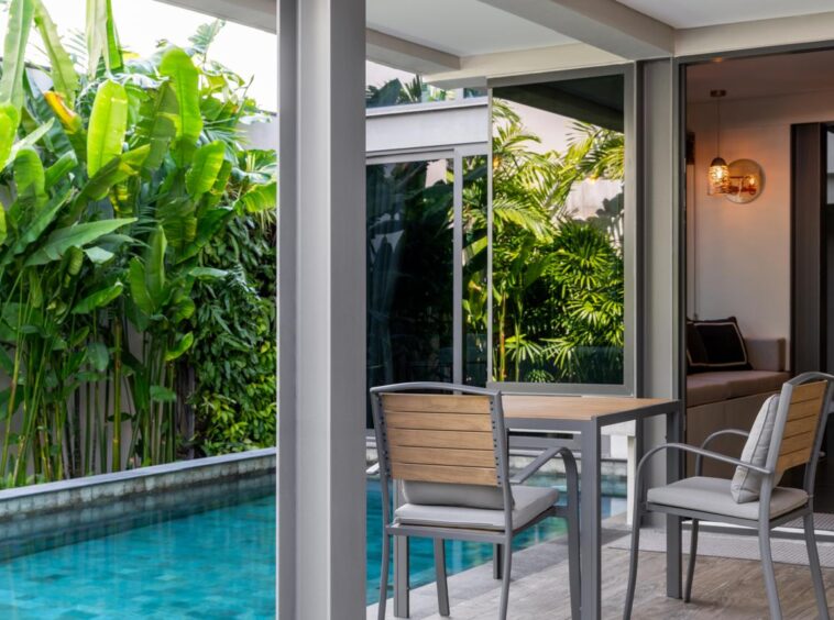 Outdoor patio with a wooden table and metal chairs beside a narrow pool, tropical plants on the side.