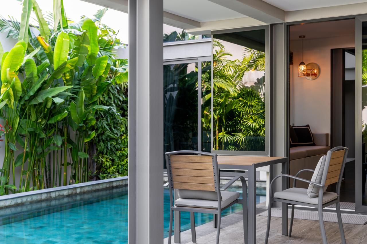 Outdoor patio with a wooden table and metal chairs beside a narrow pool, tropical plants on the side.