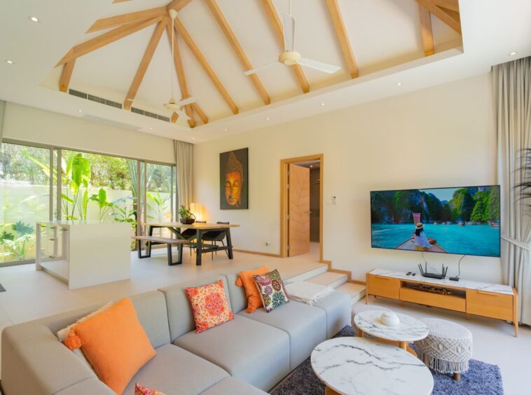 Bright open-plan living room with a vaulted wooden beam ceiling, gray sofa with orange cushions, marble-topped coffee tables, and a wall-mounted TV.