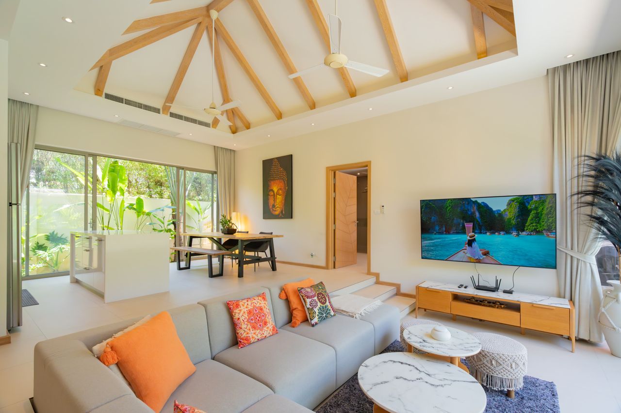 Bright open-plan living room with a vaulted wooden beam ceiling, gray sofa with orange cushions, marble-topped coffee tables, and a wall-mounted TV.