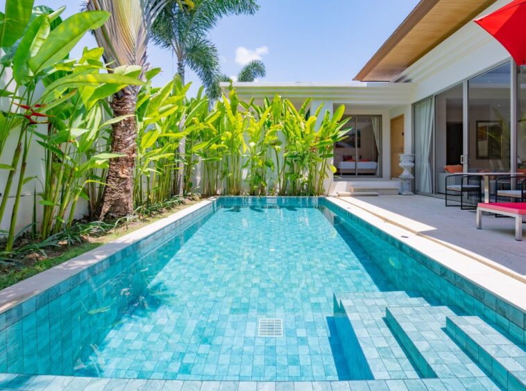 Luxurious backyard with a turquoise tiled pool, palm plants along a white wall, and a modern patio with seating and a red umbrella.