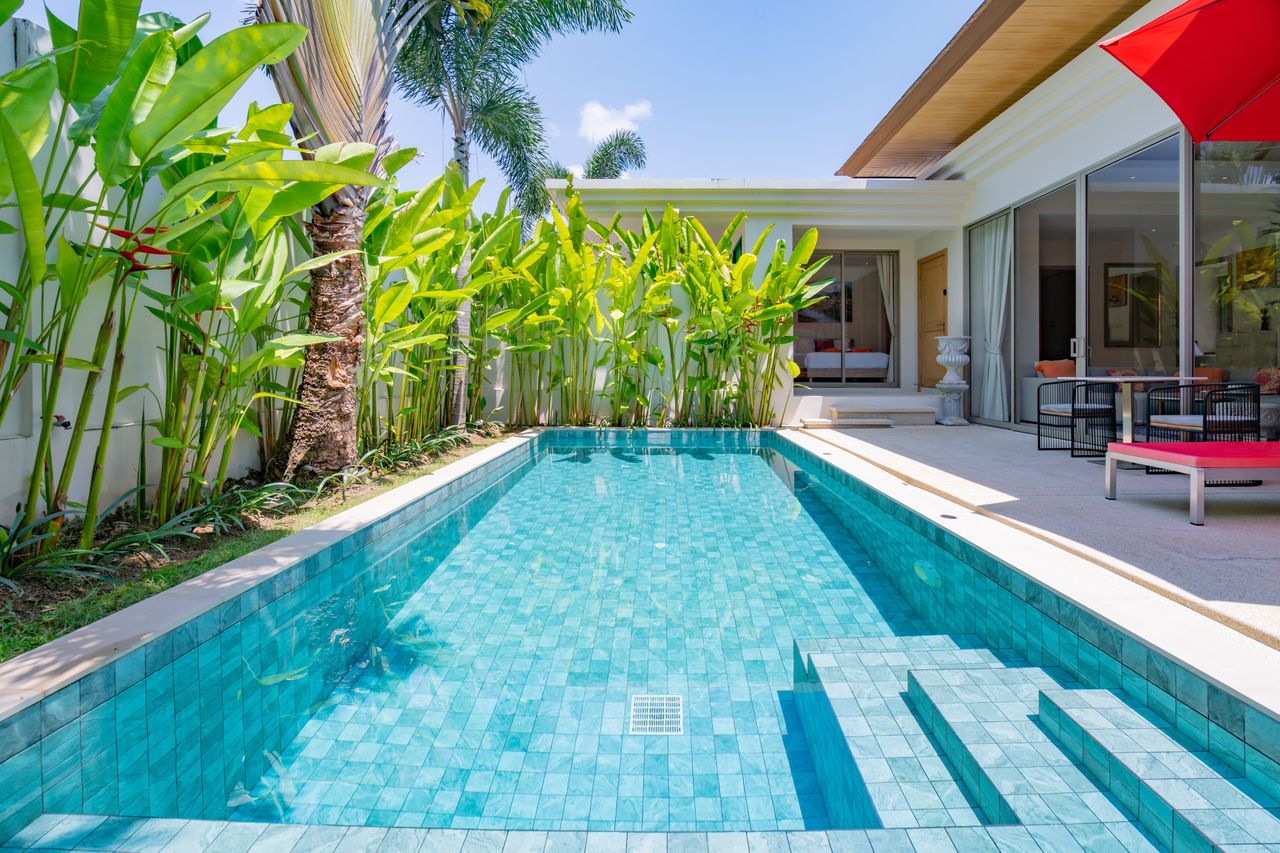 Luxurious backyard with a turquoise tiled pool, palm plants along a white wall, and a modern patio with seating and a red umbrella.
