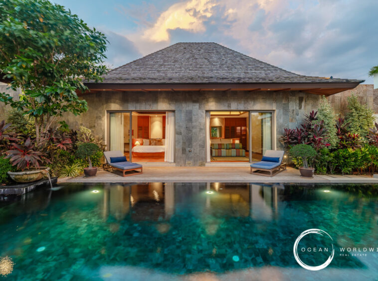Tropical villa with a stone facade, open glass doors to bedrooms/living area, lush plants, and a reflecting pool in front.