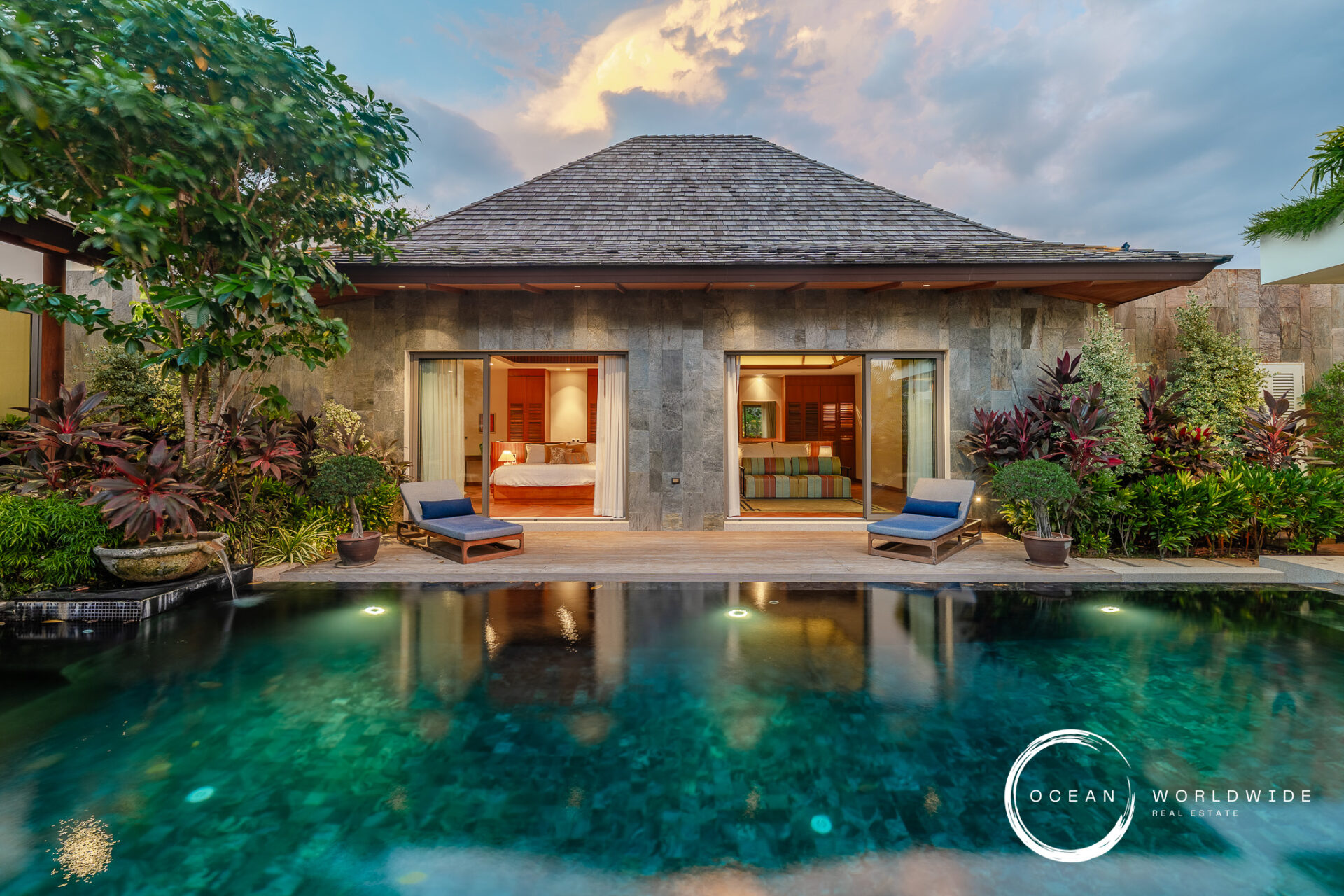 Tropical villa with a stone facade, open glass doors to bedrooms/living area, lush plants, and a reflecting pool in front.