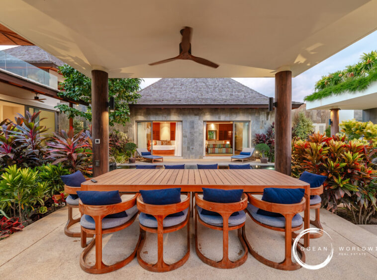 Outdoor dining area with a long wooden table and curved blue-cushioned chairs, surrounded by tropical plants in a villa courtyard under a covered patio.