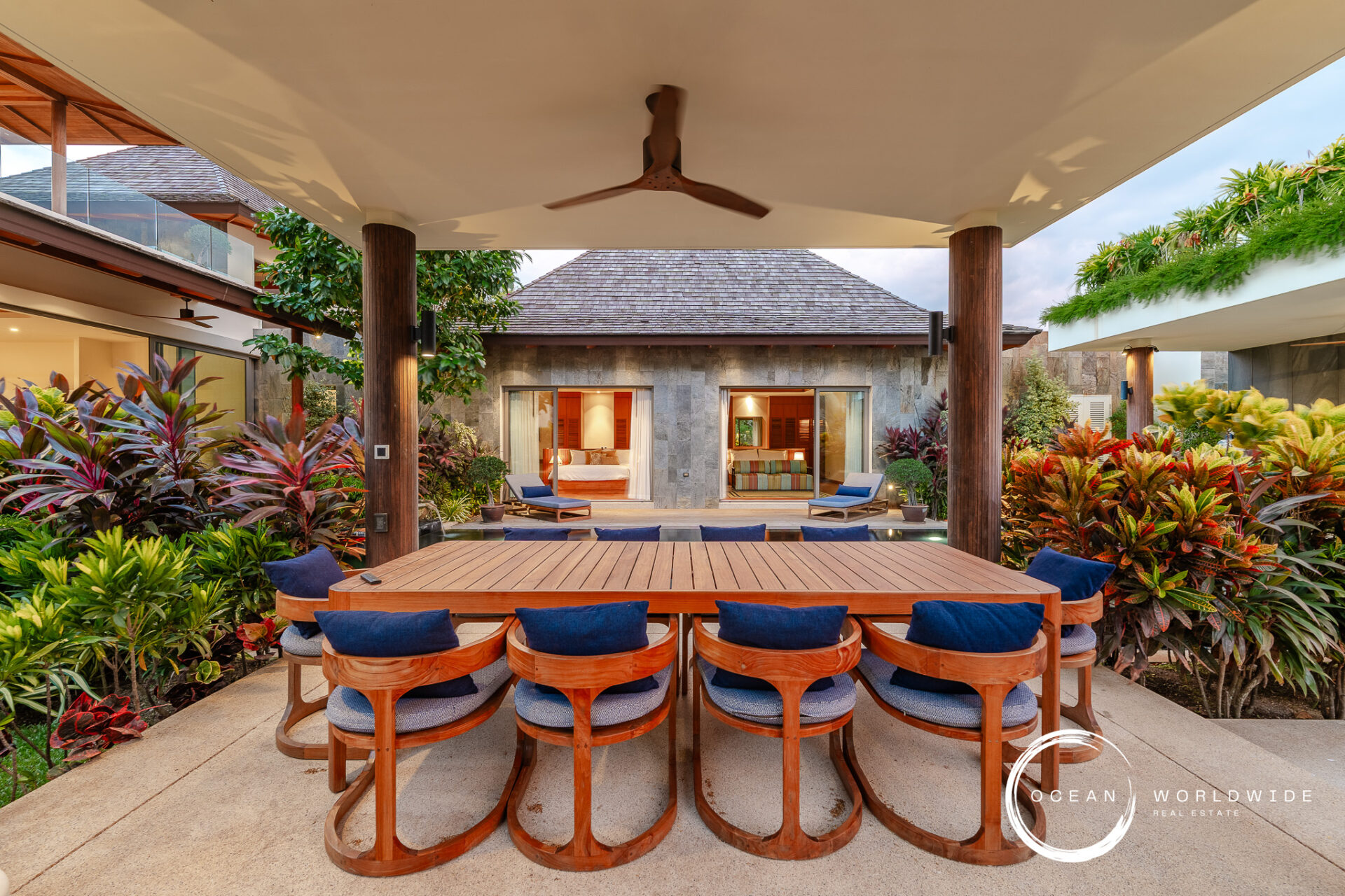 Outdoor dining area with a long wooden table and curved blue-cushioned chairs, surrounded by tropical plants in a villa courtyard under a covered patio.