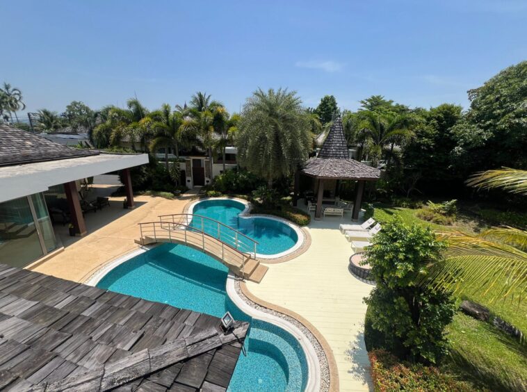 Overhead view of a tropical backyard with a curved blue pool, beige deck, and a thatched gazebo among palm trees.