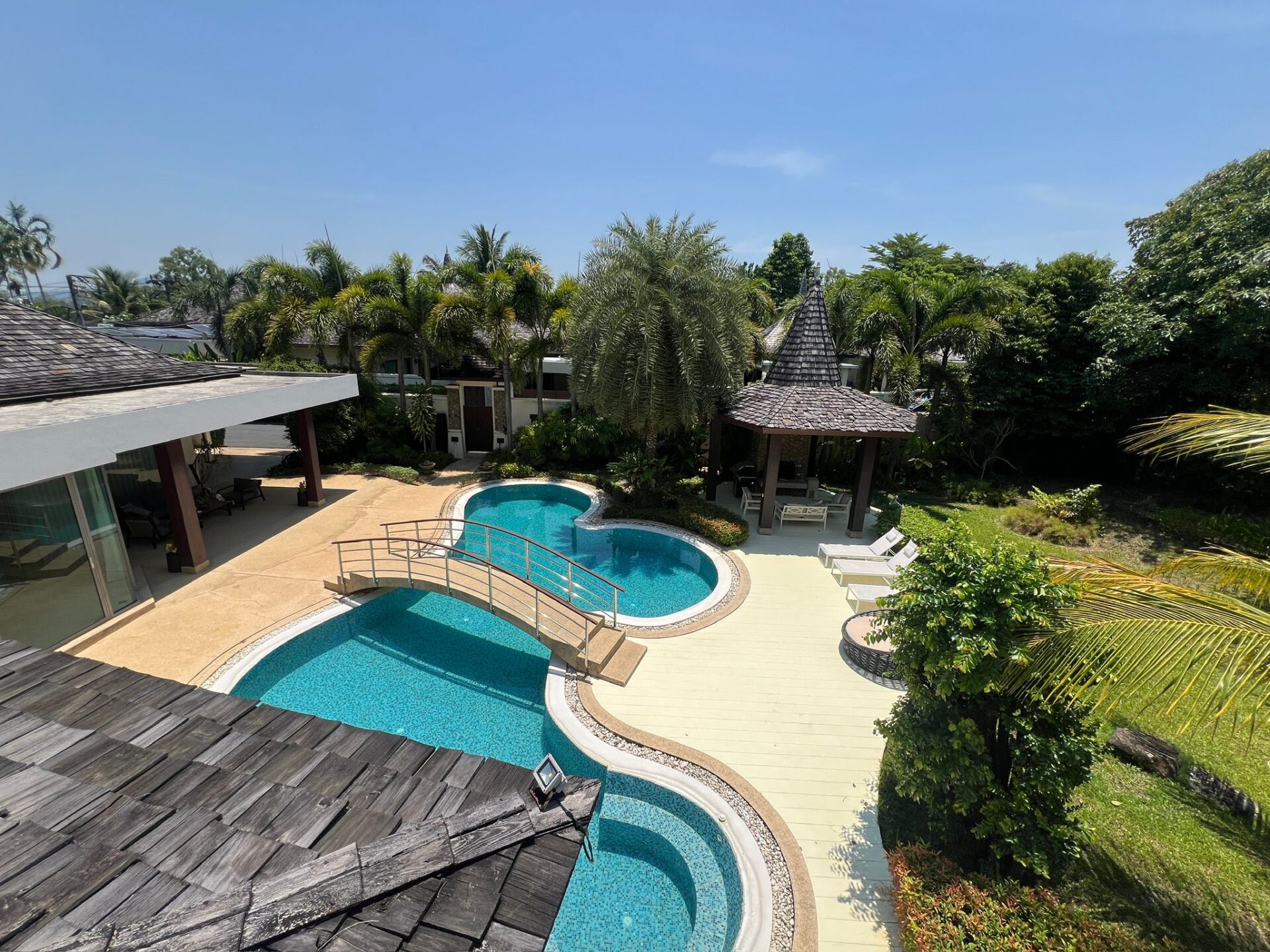 Overhead view of a tropical backyard with a curved blue pool, beige deck, and a thatched gazebo among palm trees.