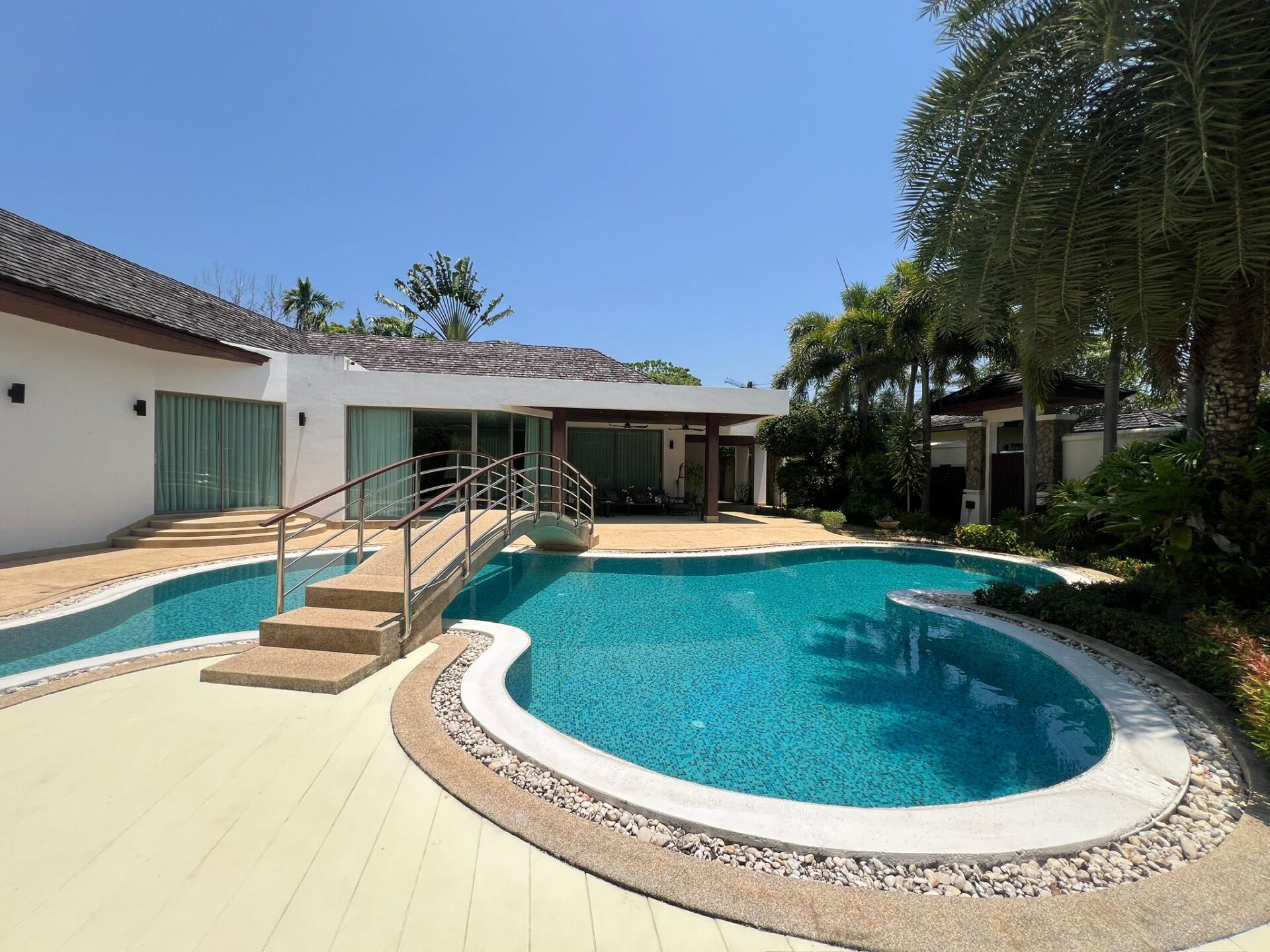 Outdoor backyard with a curved pool, tile steps, and a beige deck beside a white building under a blue sky. Palm trees frame the scene.
