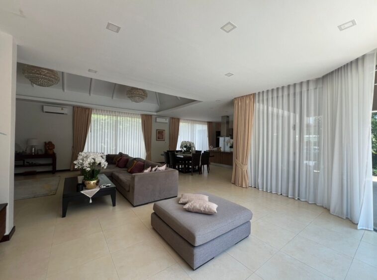 Spacious, modern living room with gray sectional sofa, glass coffee table, and white tiled floor; large windows with sheer curtains let in natural light.